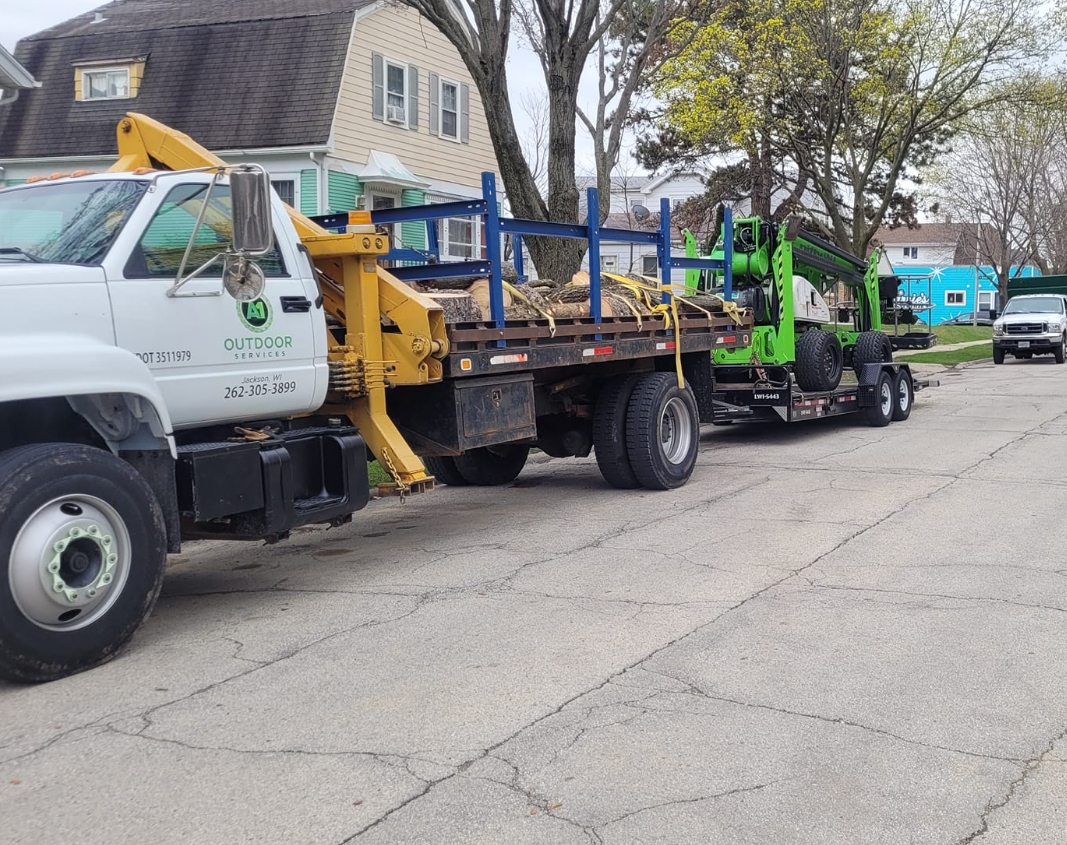 White truck towing a flatbed trailer loaded with logs and a green wood chipper parked on a street.