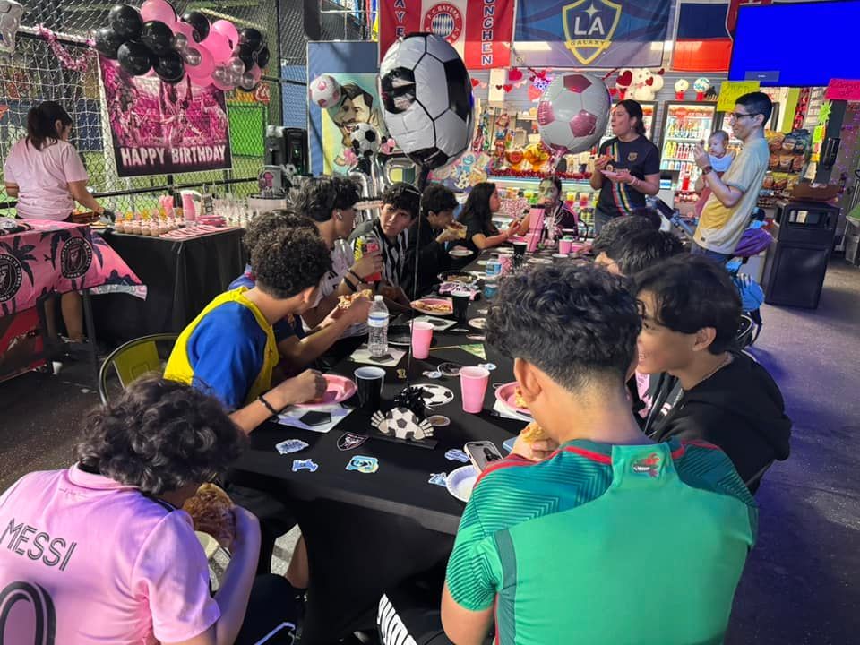 People at a soccer-themed party, sitting around a table with decorations, balloons, and food.