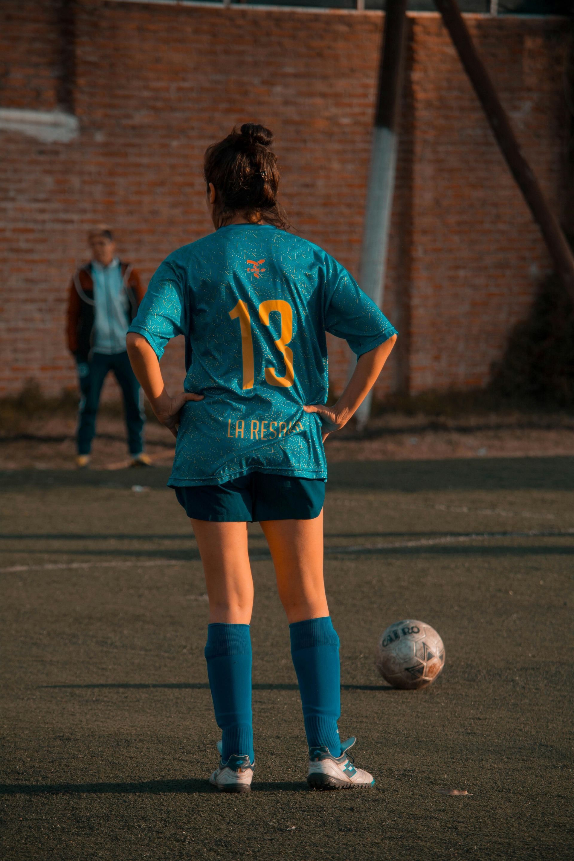 Soccer player in teal jersey with number 18, hands on hips, on field with ball; person in background.