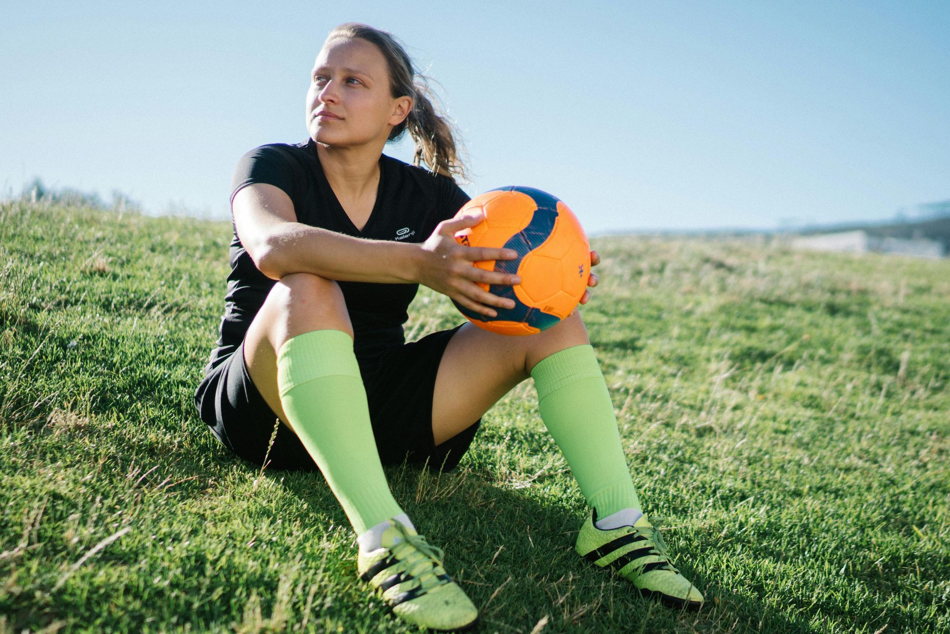 Woman in soccer gear sits on grass, holding a ball. Green socks and cleats, black shirt and shorts. Sunny outdoor setting.
