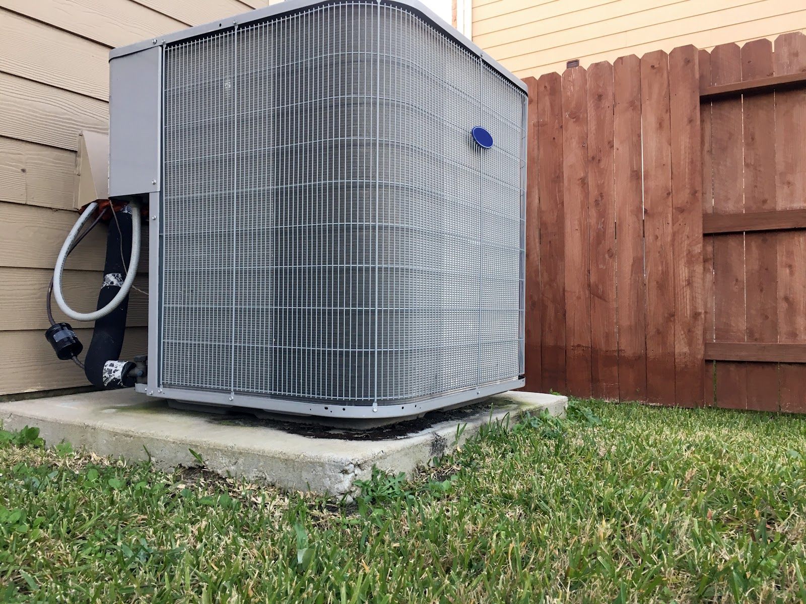 Air conditioning unit on a concrete pad next to a wooden fence and grass lawn.