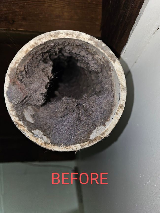 Close-up view of a dusty, clogged dryer vent. The vent is white, filled with gray lint and debris.