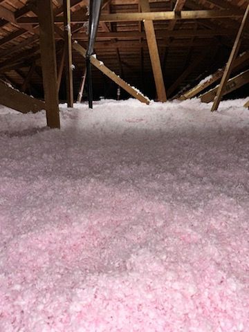 Pink insulation fills an attic space, with wooden beams and rafters overhead.