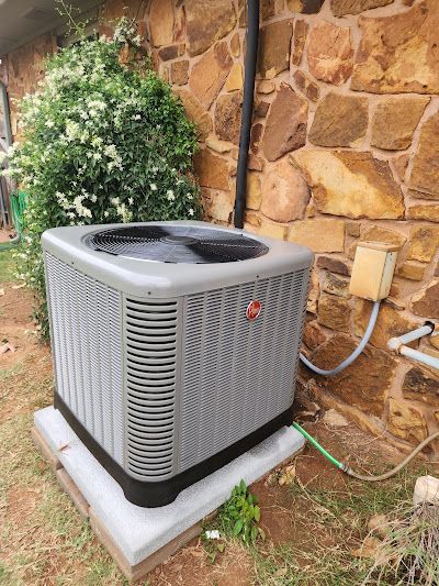 An air conditioning unit next to a stone wall with a bush behind it.