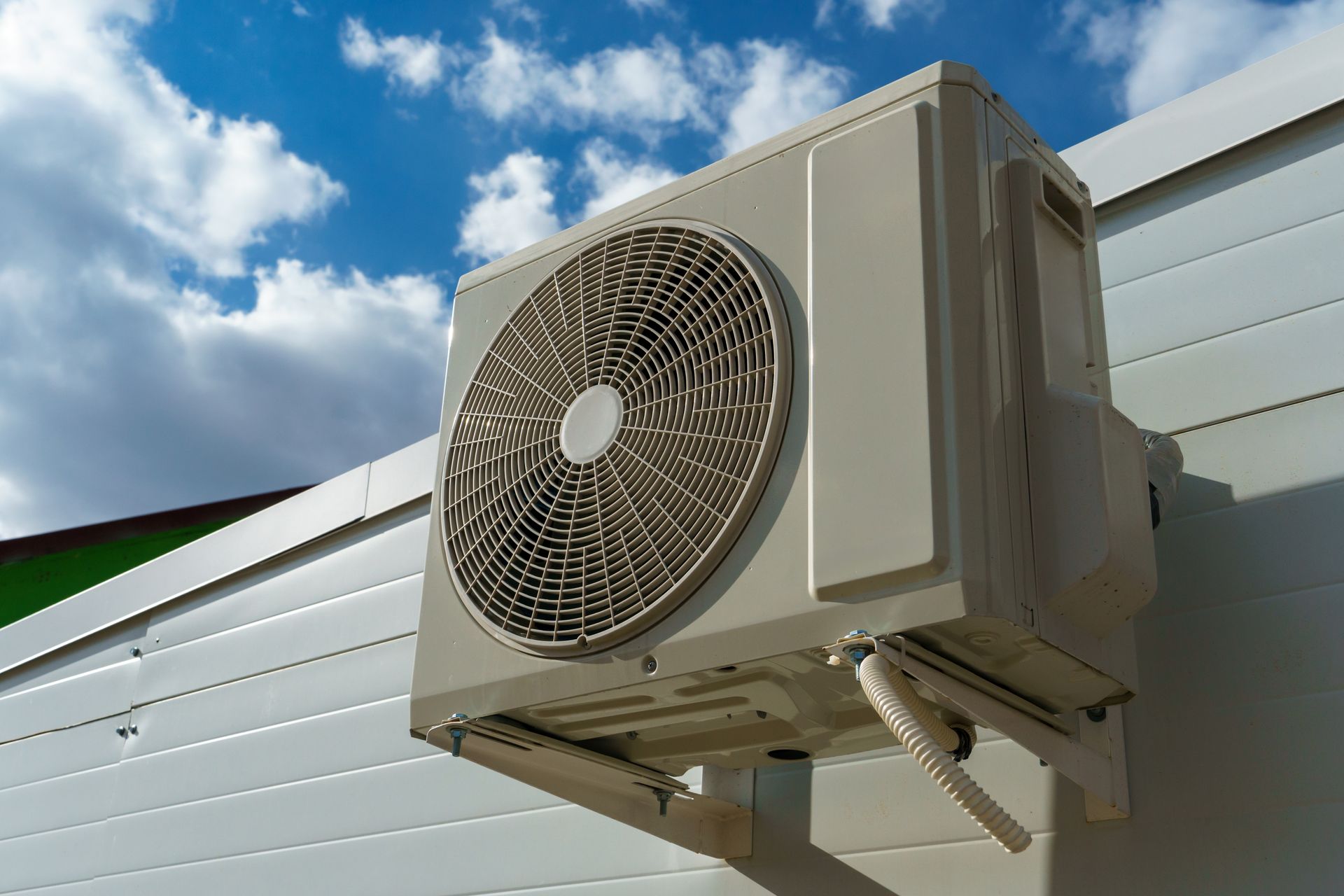 Exterior air conditioning unit mounted on a white wall under a partly cloudy blue sky.