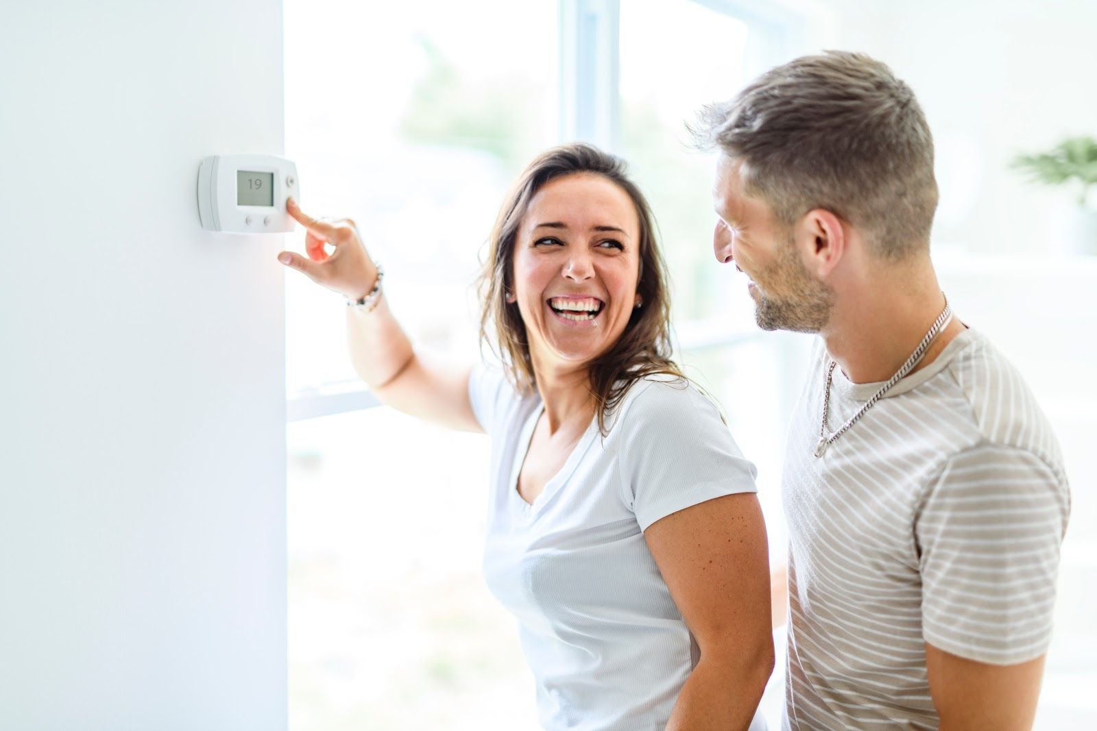 Woman adjusting a thermostat; smiling at a man in a white-walled room with natural light.