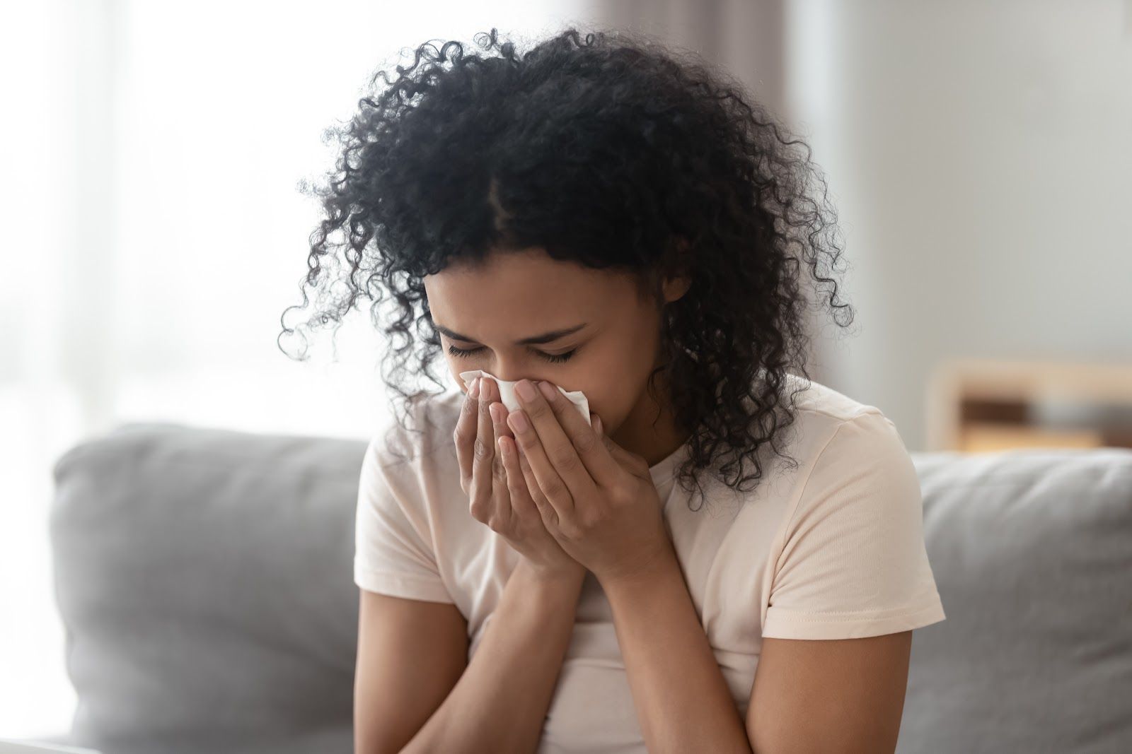 Woman sneezing into a tissue on a gray couch; white shirt, curly dark hair.