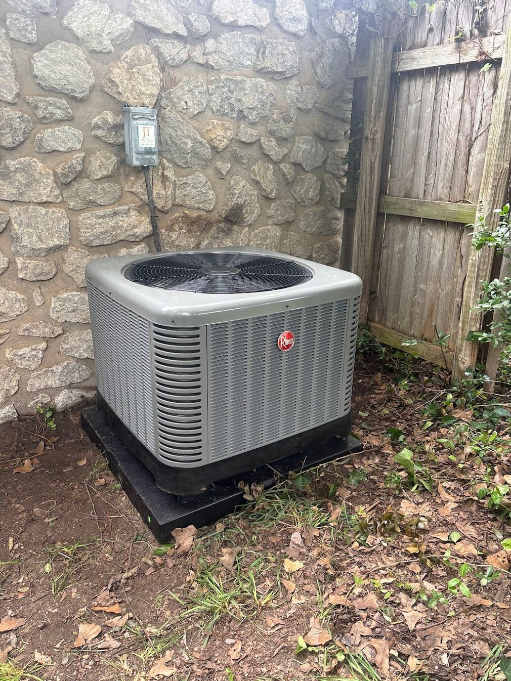 Gray air conditioning unit next to a stone wall and wooden fence.