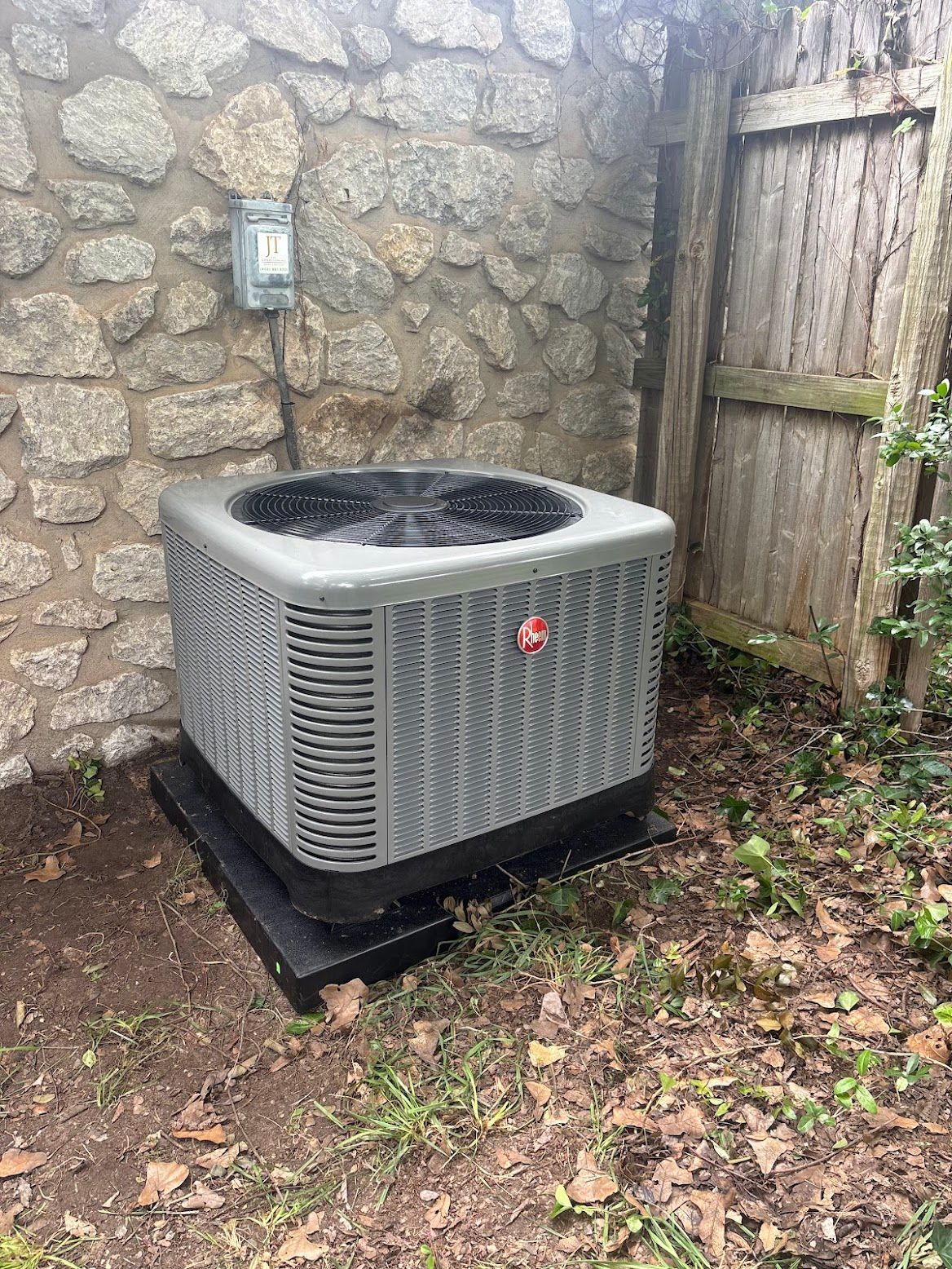 Gray air conditioning unit next to a stone wall and wooden fence.