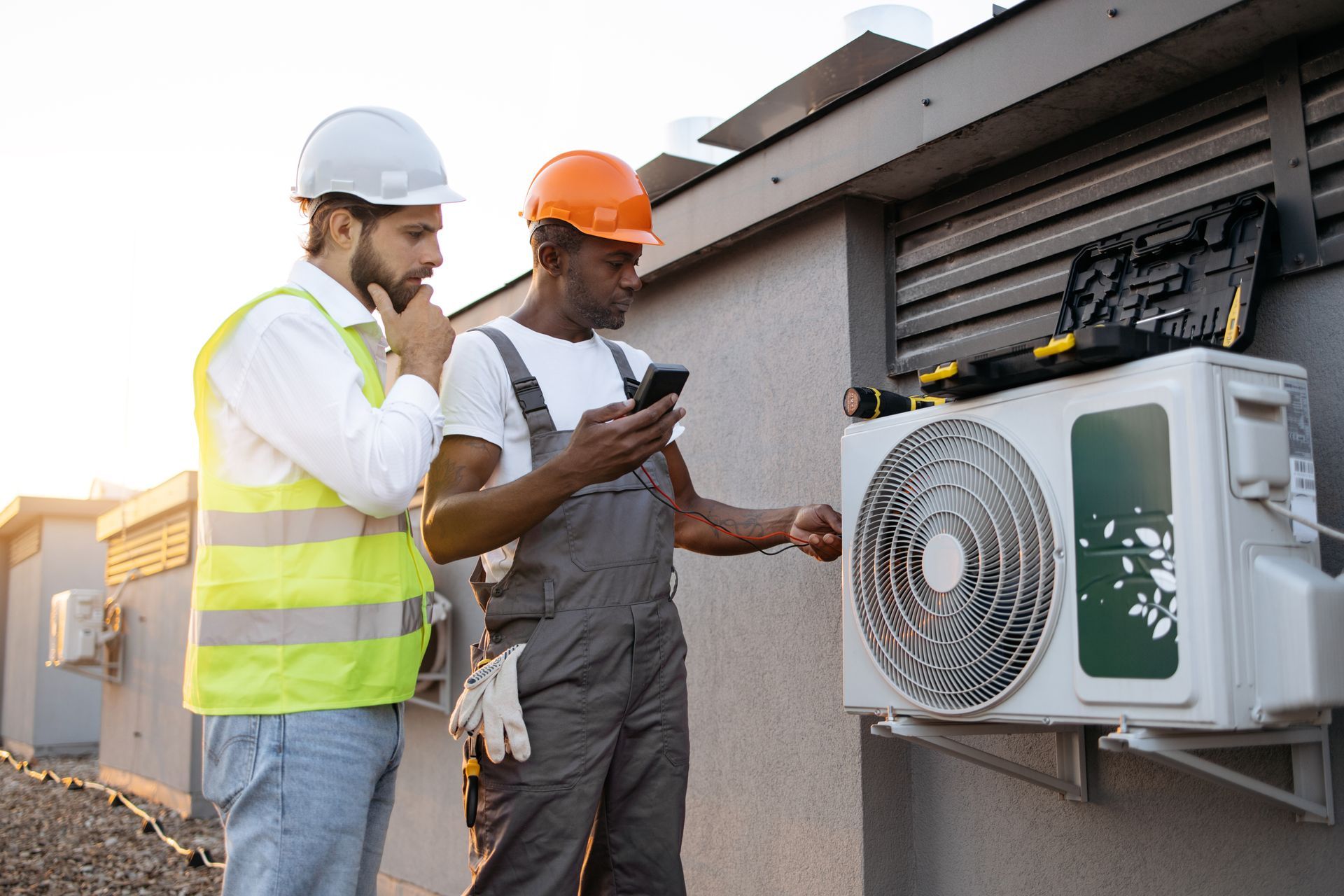 Two workers examine an AC unit, one using a phone, the other in thought.