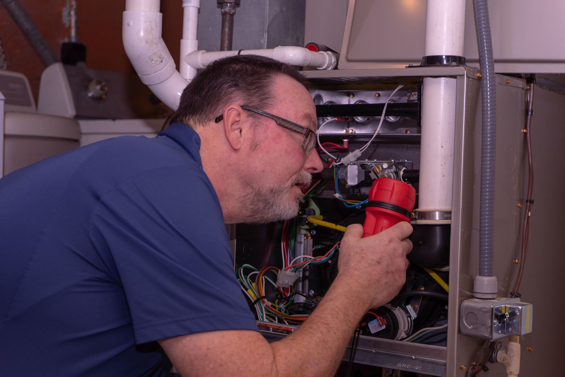 Technician examines furnace interior with a flashlight in a utility room.