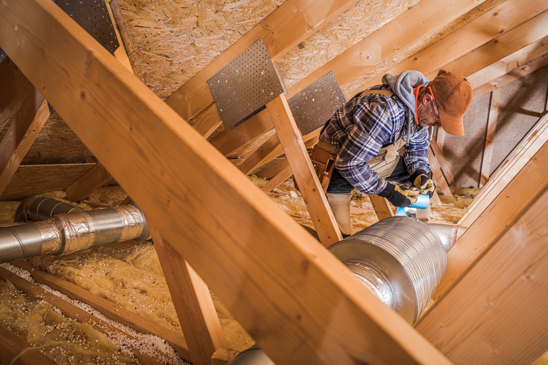 A person in an attic is working with ductwork and insulation, wearing a cap and safety glasses.