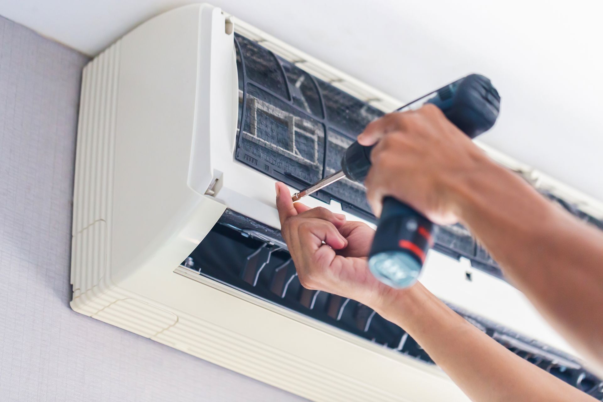Person using a power drill to repair a white wall-mounted air conditioning unit.