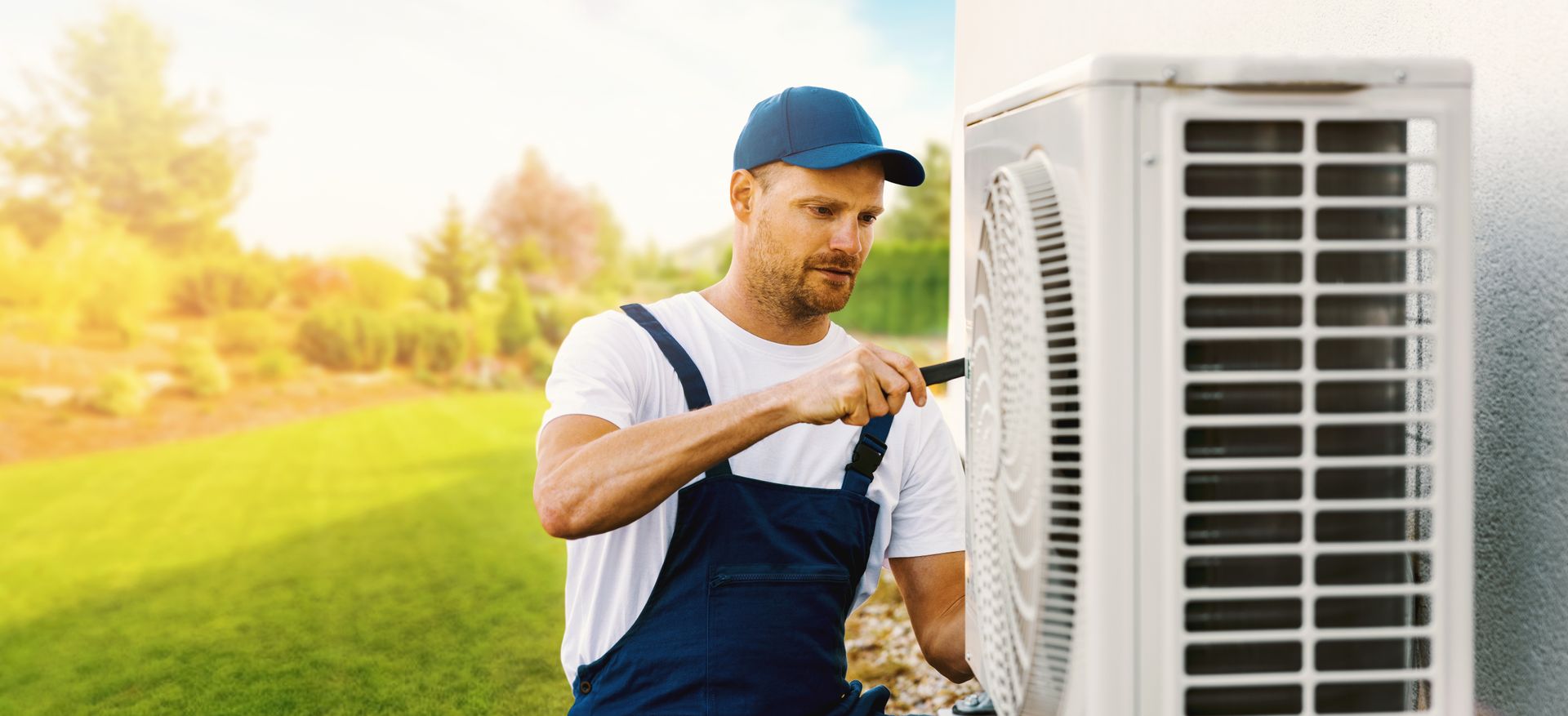 HVAC technician in a blue uniform, fixing an outdoor air conditioner unit in a sunny yard.