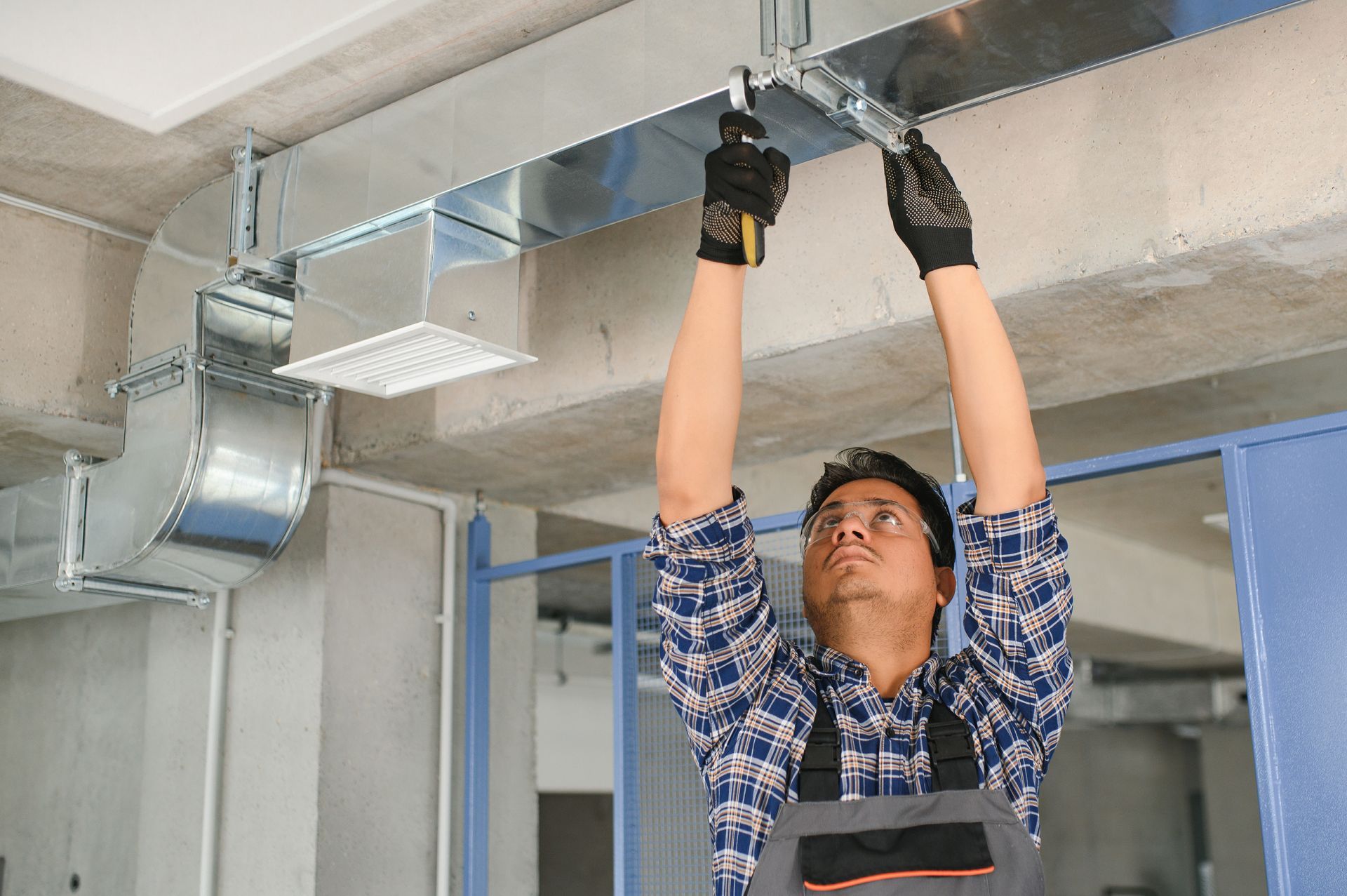 Man installing HVAC ductwork in a building, wearing gloves and overalls.