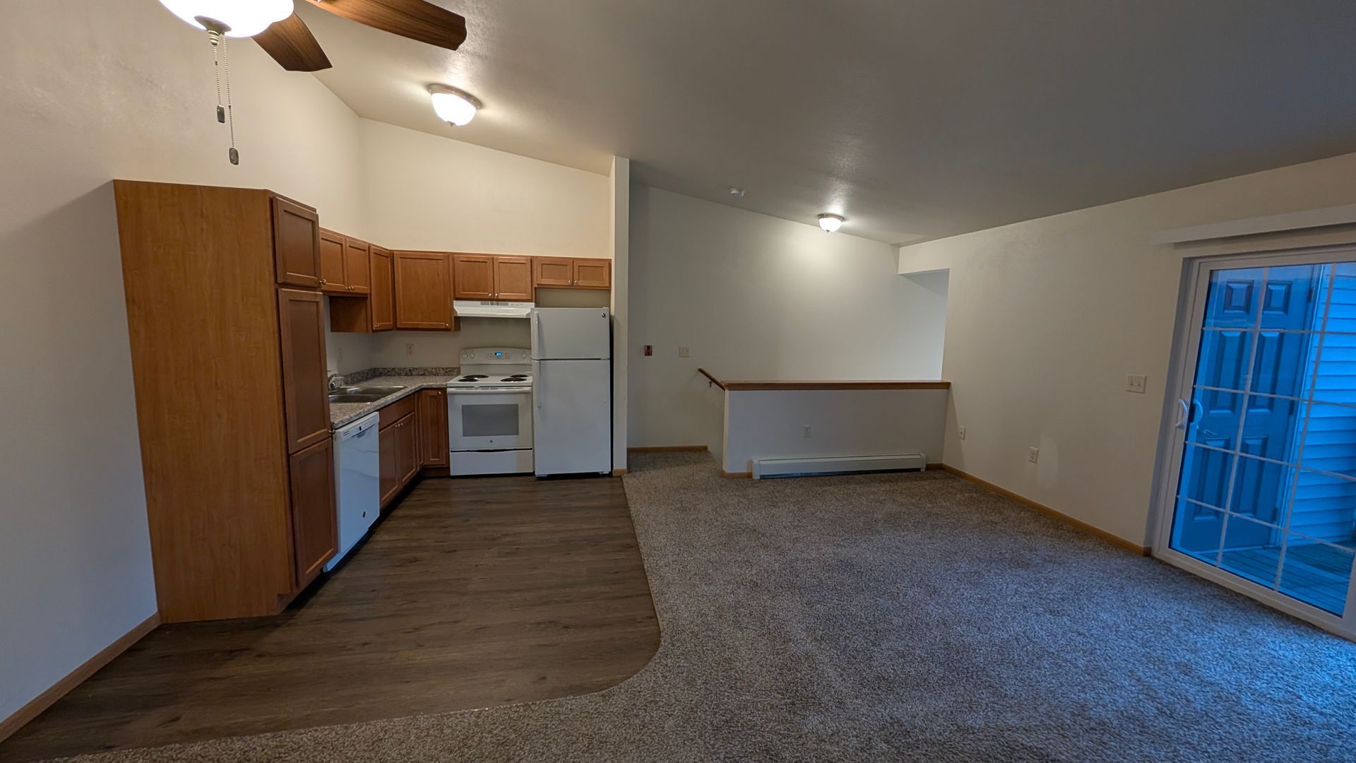 Empty apartment living area with kitchen cabinets, white appliances, carpet, and blue sliding glass door