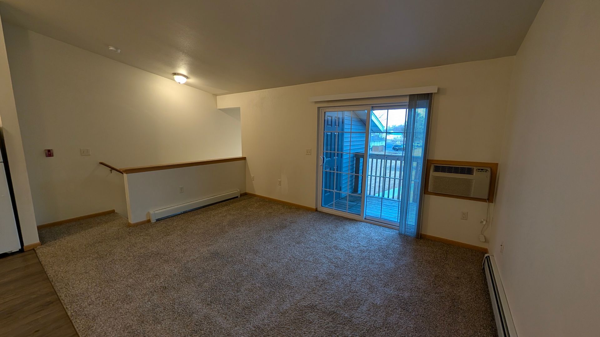 Empty carpeted living room with sloped ceiling, wall light, and sliding glass doors to a balcony.