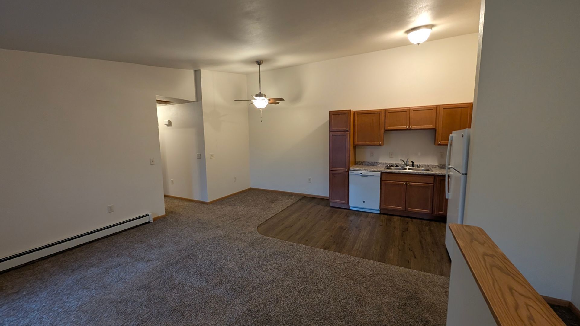 Empty living room with carpet, ceiling fan, and small kitchen area with wood cabinets.