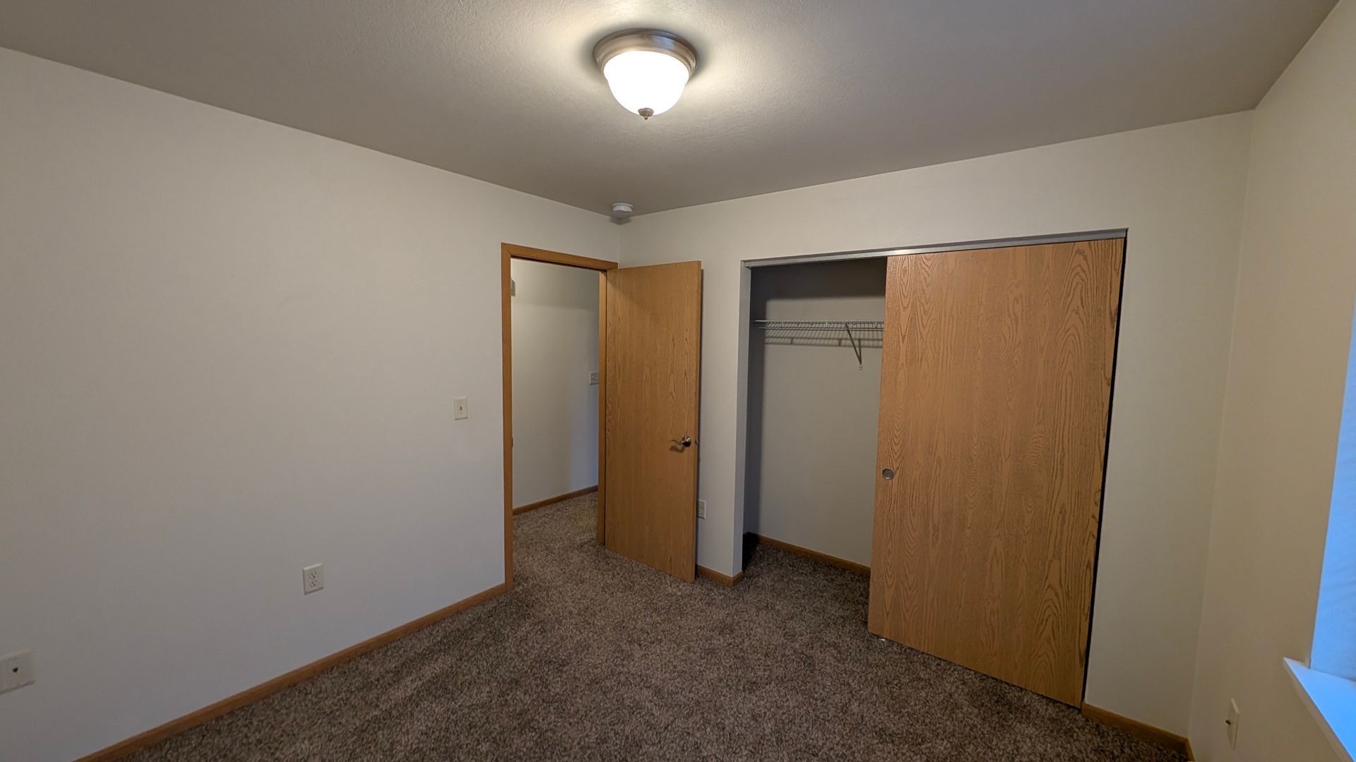 Empty beige bedroom with open closet doors and carpeted floor, lit by a ceiling light.