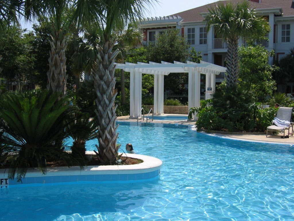 Swimming pool with white pergola, palm trees, and a building in the background.
