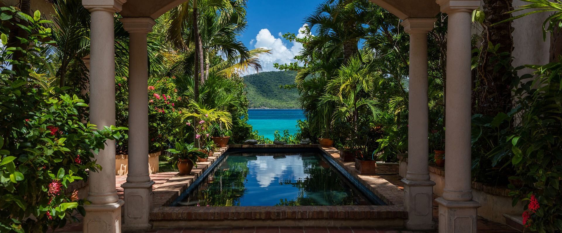 View of a turquoise ocean and sky through a garden with a reflecting pool.