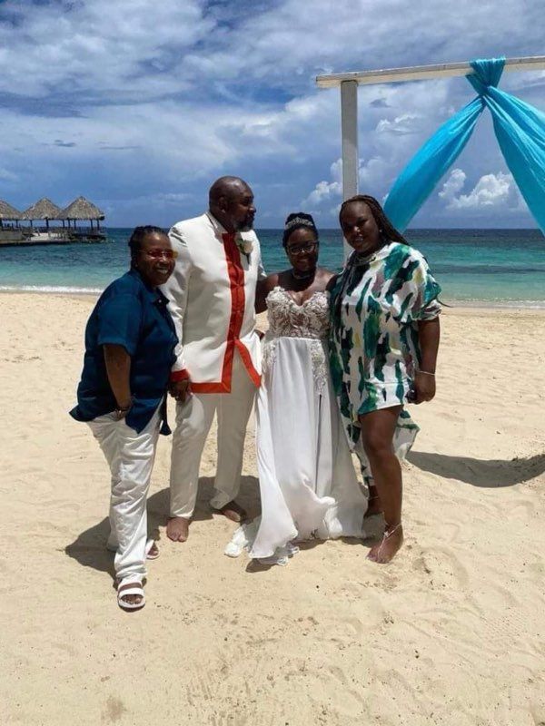 Wedding group on beach: Bride in white, groom in white with red trim. Guests stand beside them. Blue sky.