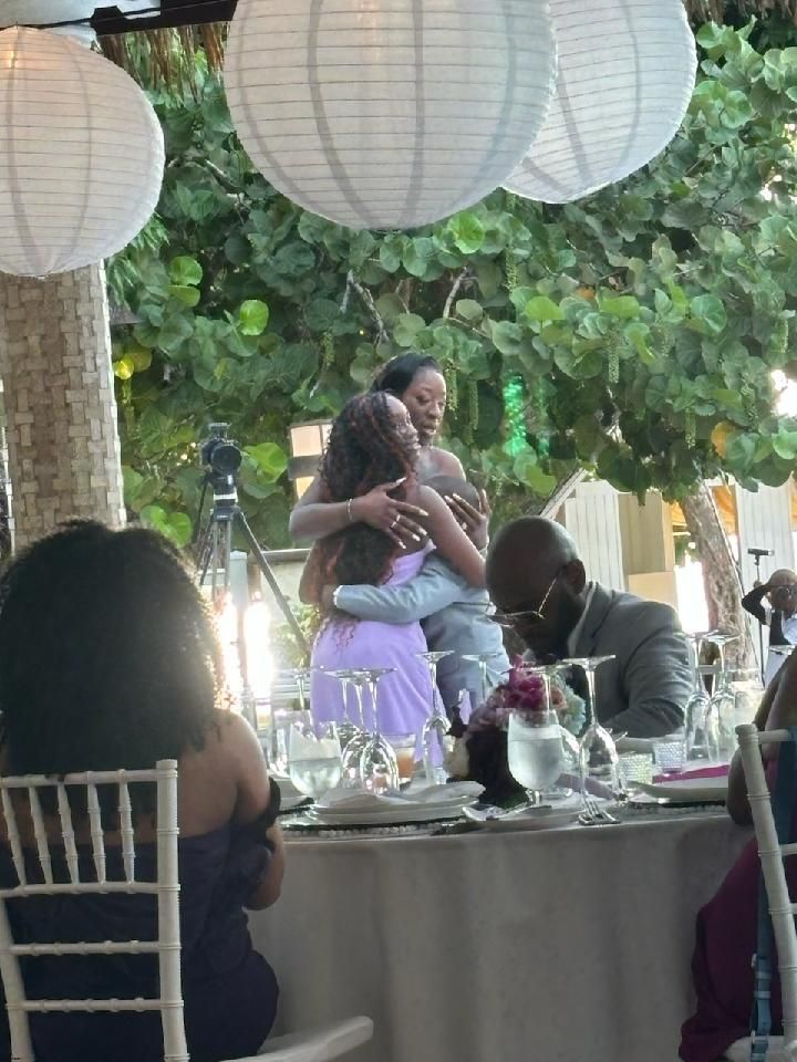 People hugging at a wedding reception with white lanterns, a seated guest, and a table setting.