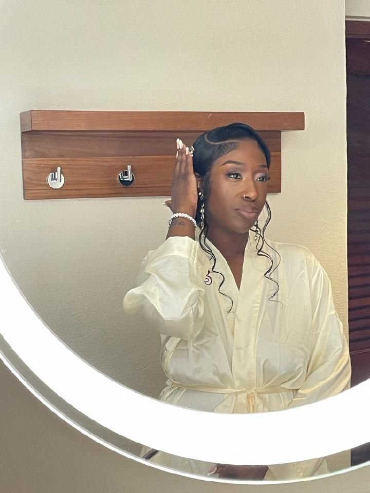 Woman in a cream robe looking in a circular mirror, hand in hair. White walls, wooden shelf.