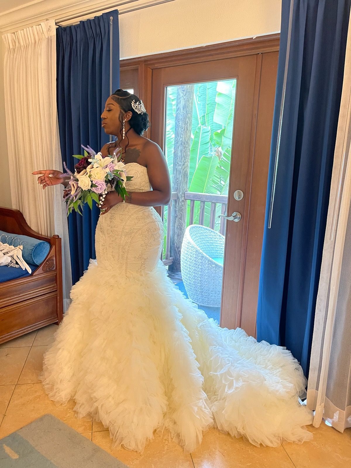 Bride in a white strapless gown, holding a bouquet, stands near blue curtains and an open door.