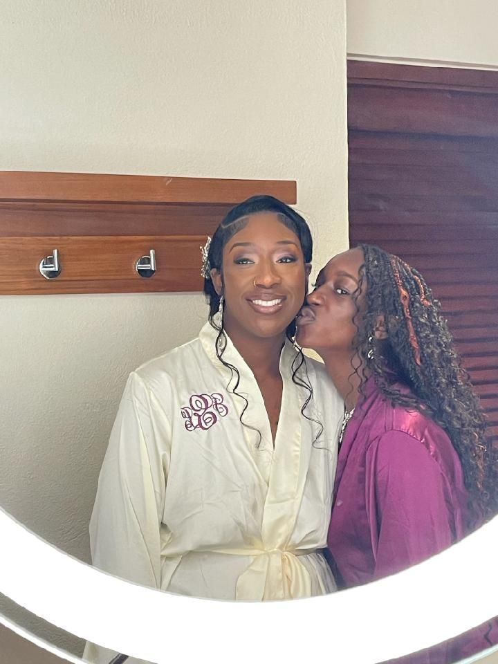 Woman in white robe smiles as woman kisses her cheek in front of a mirror.