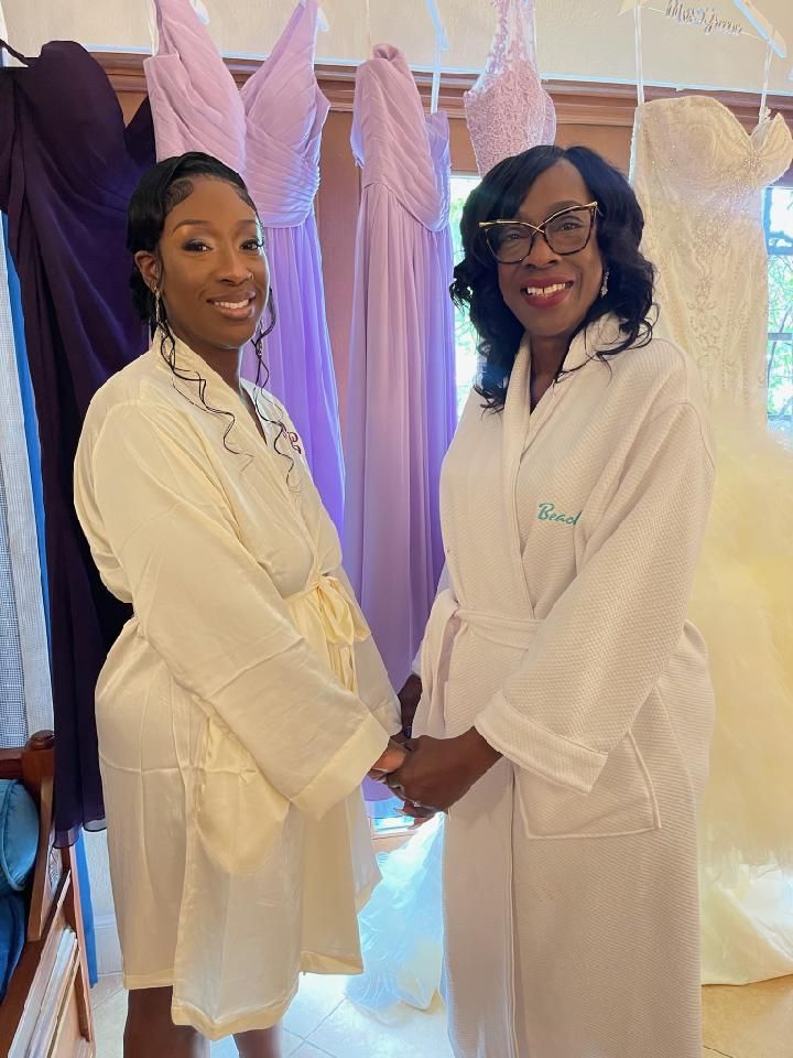 Two women in white robes holding hands, standing in front of bridesmaid dresses.