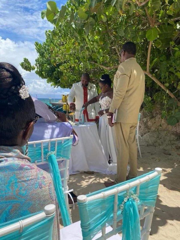 Beach wedding: Bride and groom near white table; turquoise chairs face them. Man in white coat officiates.