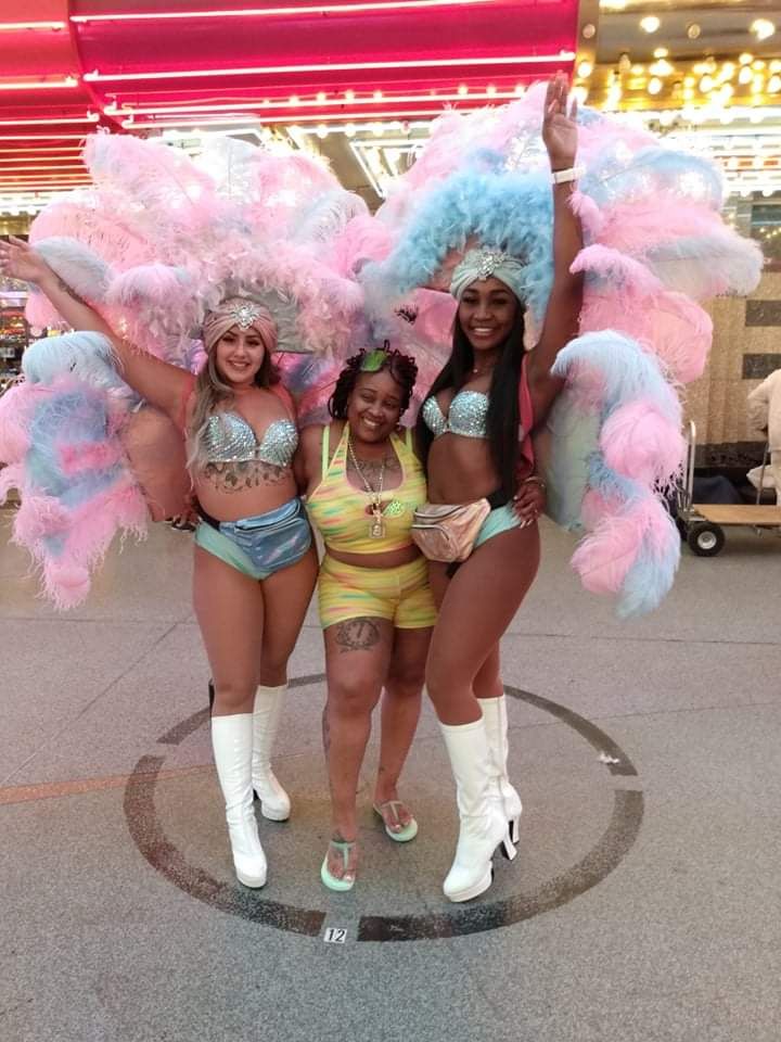 Three women pose on a city street: two in elaborate feather costumes, one smiling in yellow shorts.