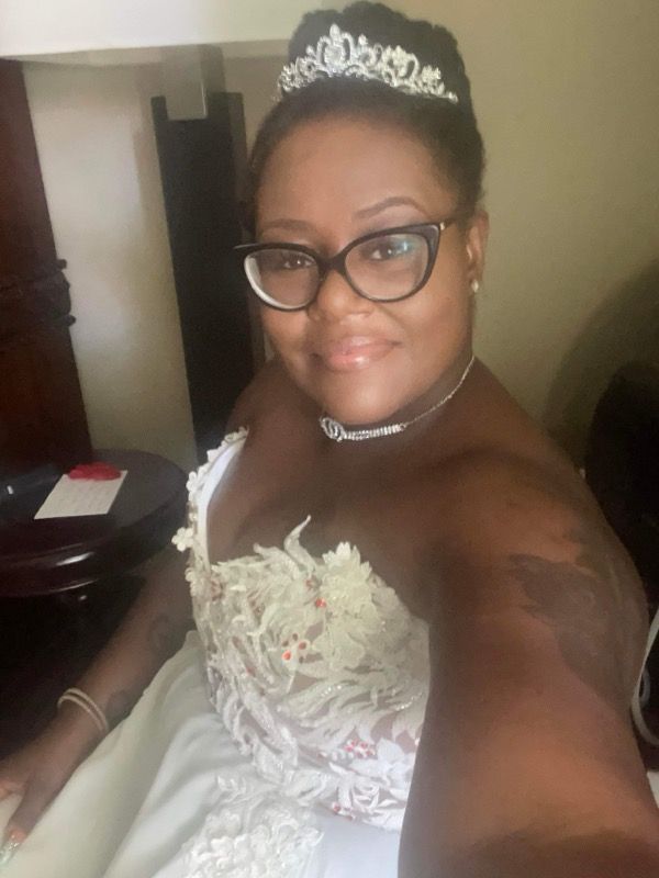 Woman in wedding dress, tiara, glasses, smiles for selfie. Inside room, small round table.