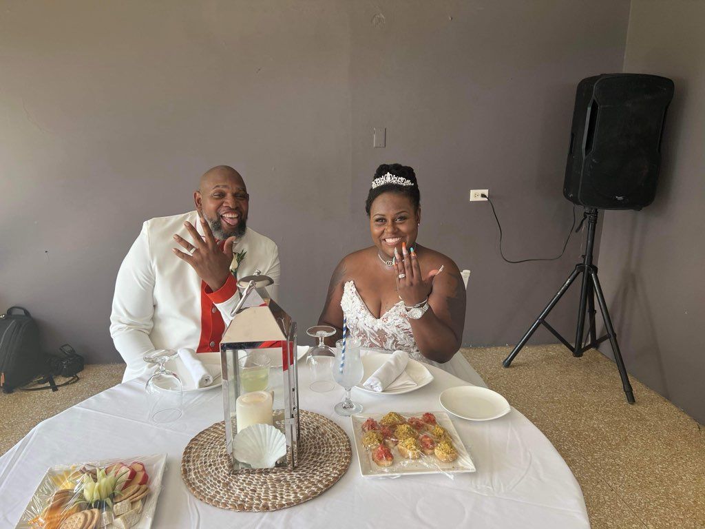 Newlyweds at a table, showing off rings. The man wears white, the woman a white dress.