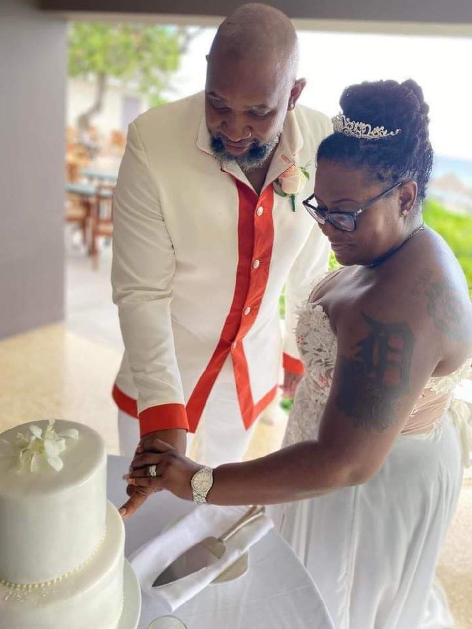 Couple cuts a wedding cake outdoors. Man in white suit with orange trim, woman in white dress, both smiling.