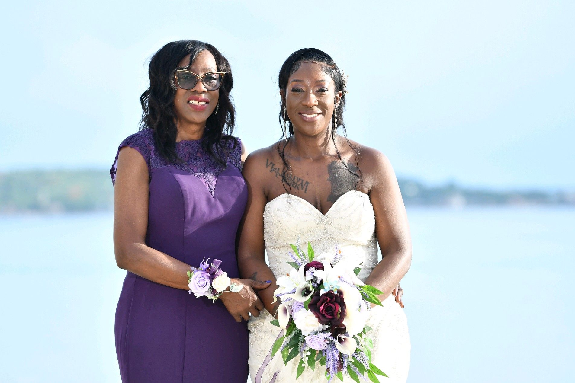 Bride and woman pose by water. Bride in white dress, holding bouquet. Woman in purple dress. Sunny day.