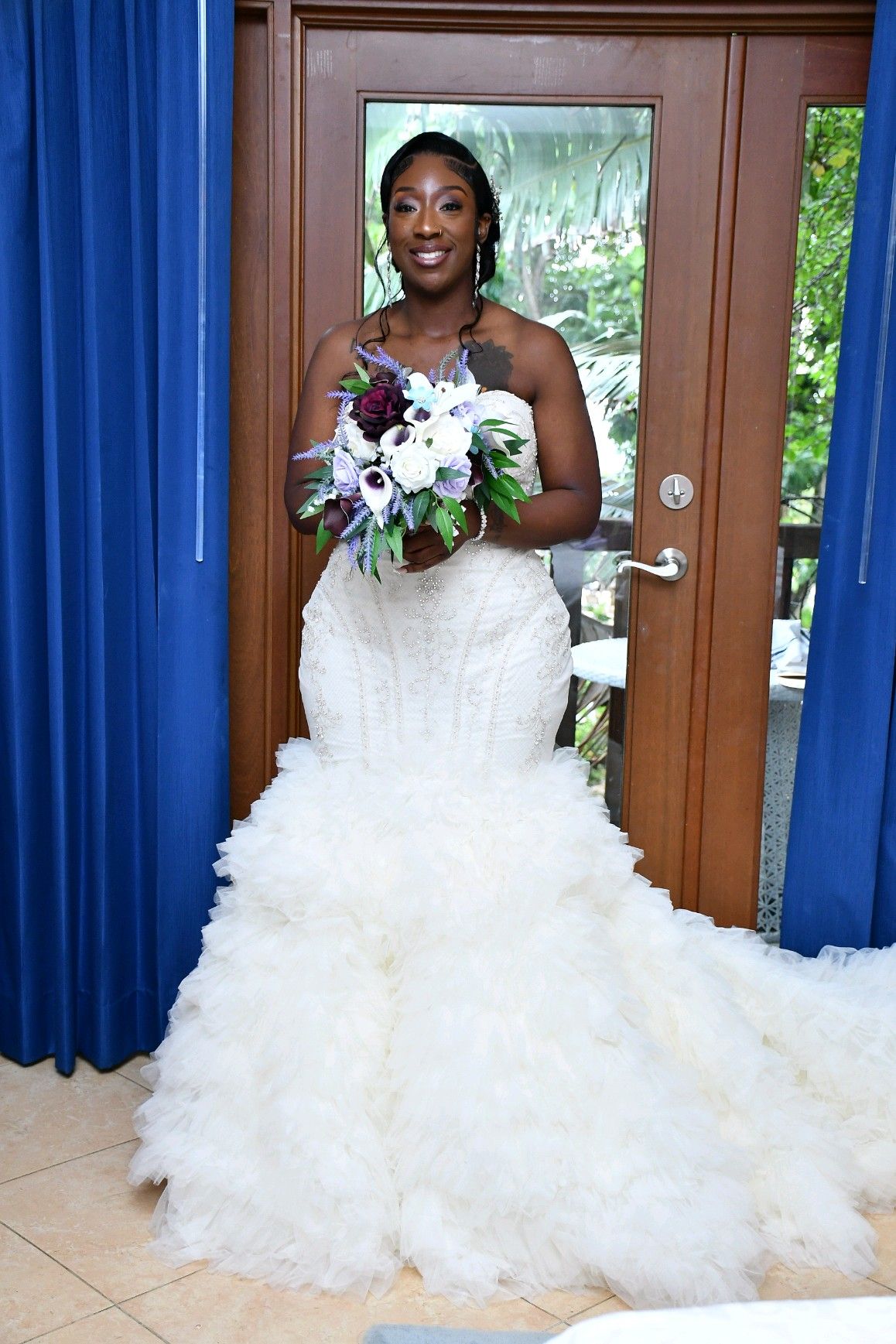Bride in white strapless mermaid gown, holding bouquet, smiles. Standing near blue curtains, doorway open to trees.
