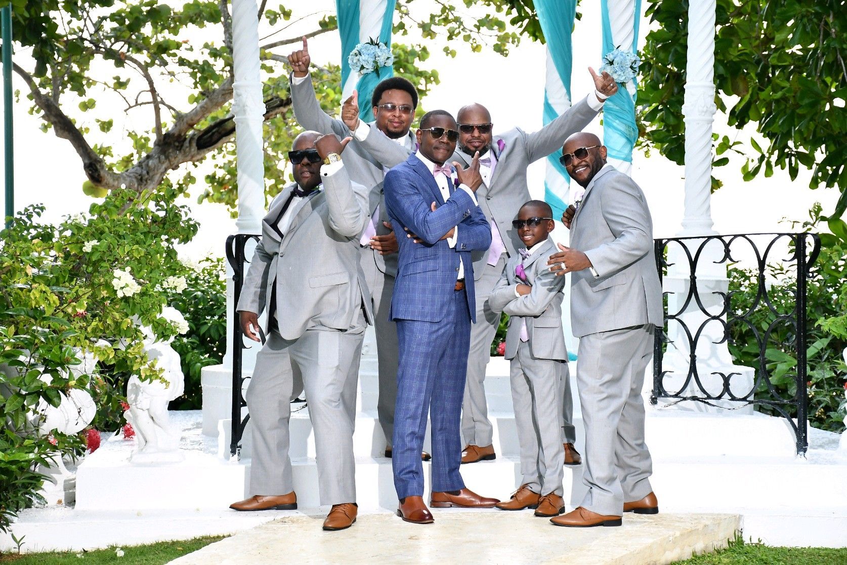 Groomsmen in gray suits pose outdoors with a man in a blue suit; all are wearing sunglasses.