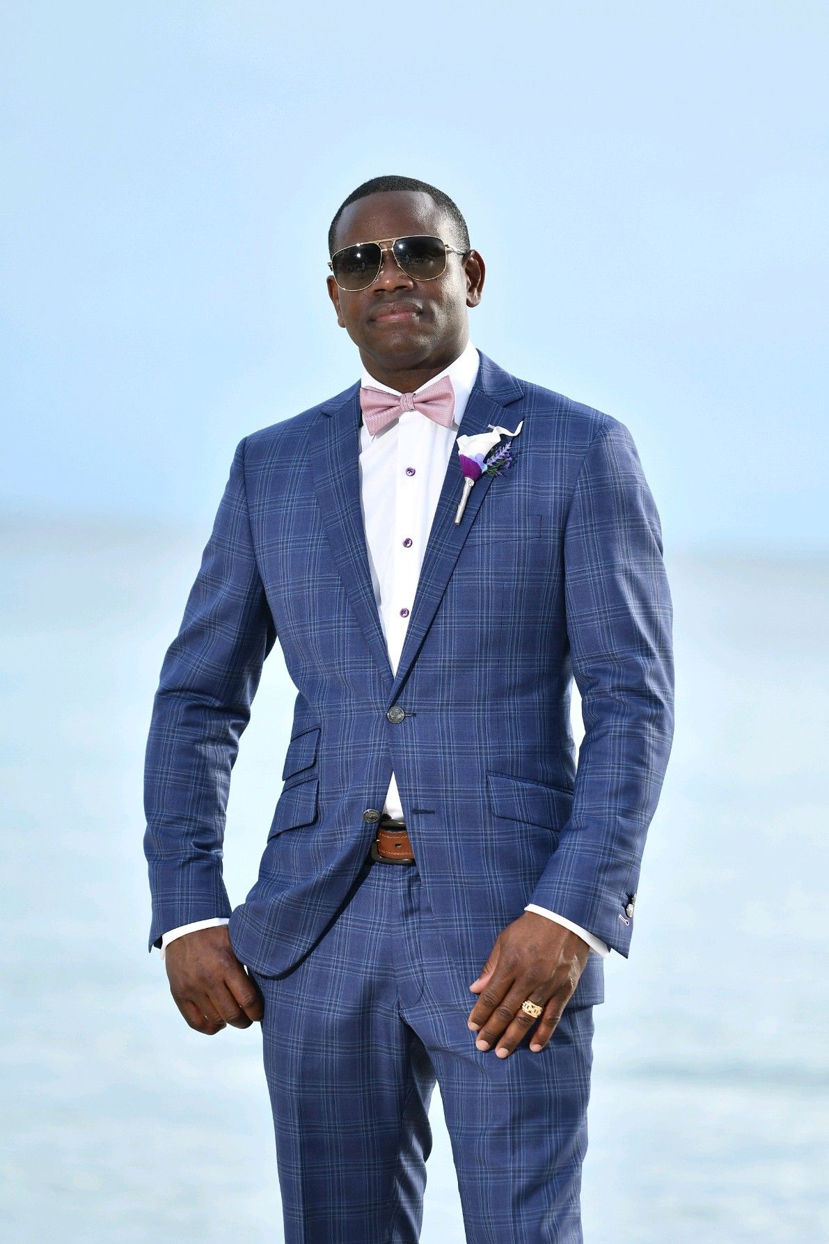 Man in blue suit and pink bow tie, sunglasses, standing near water.