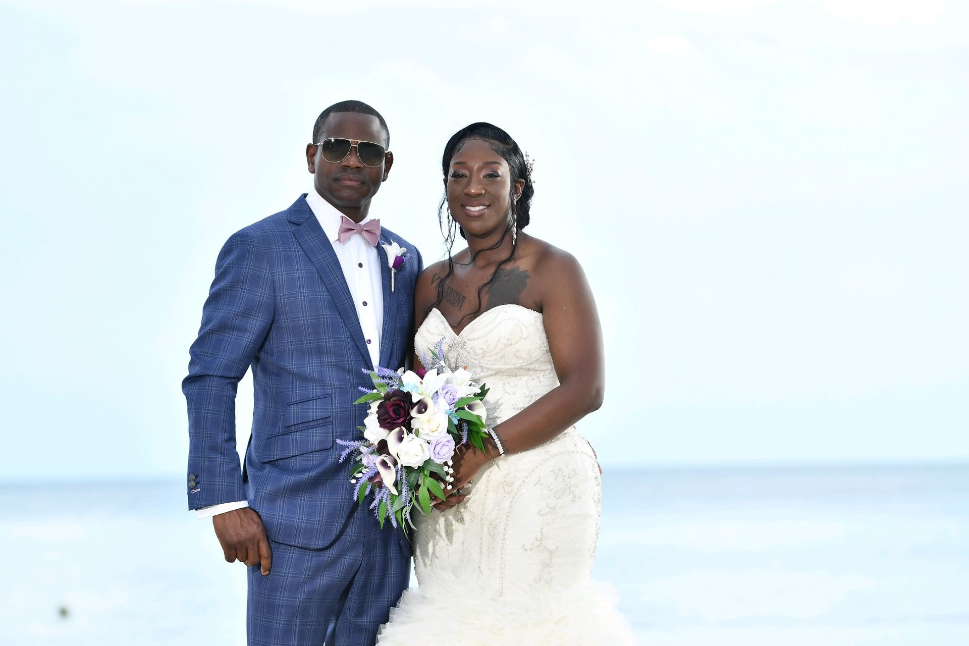 Newlyweds pose by the ocean. Man in blue suit and bow tie, woman in white strapless gown, holding bouquet.