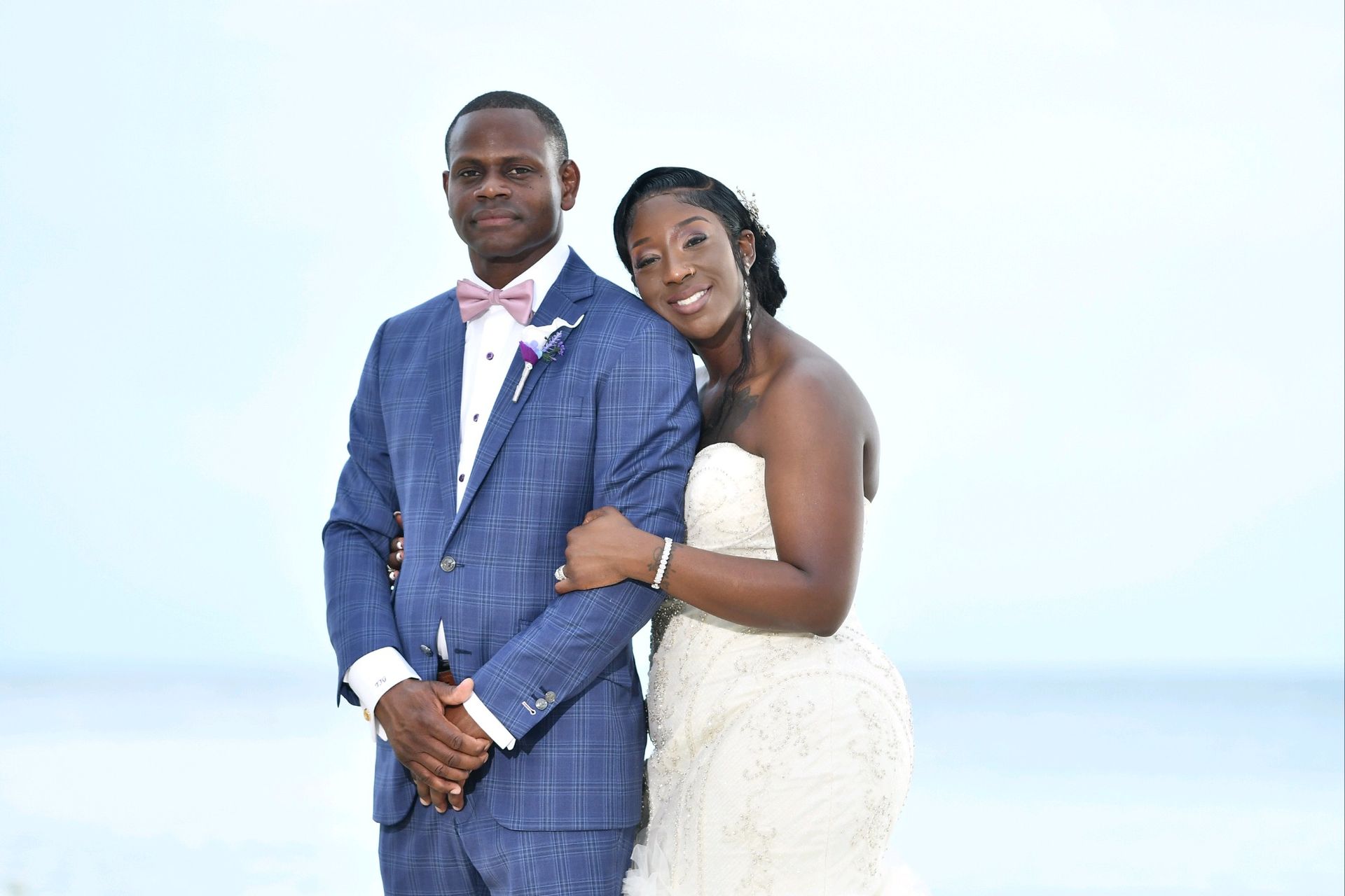 Bride and groom embrace on a beach; he in blue suit, she in a white gown, both smiling.
