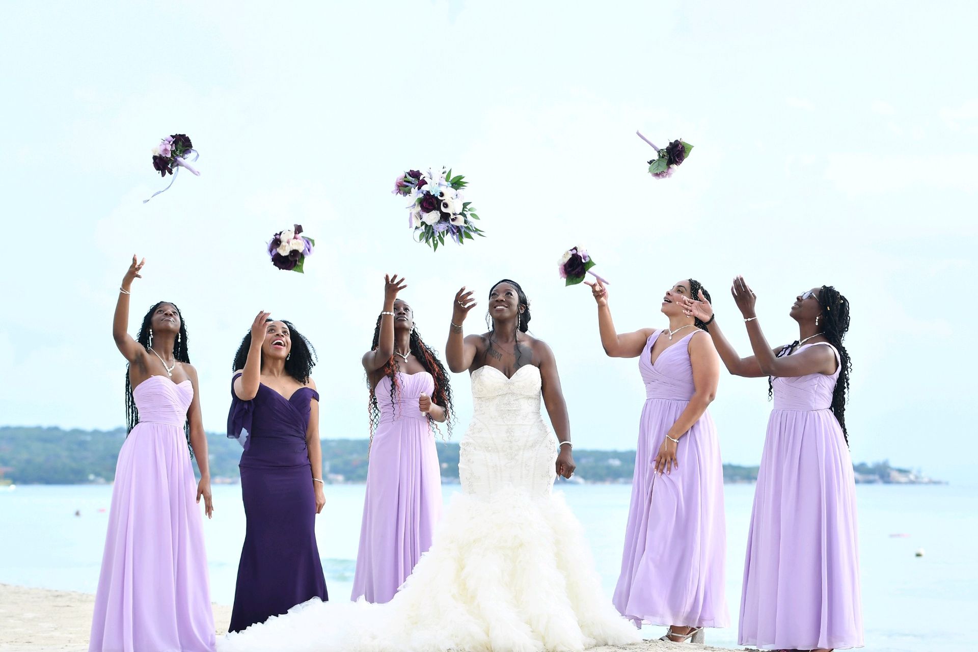 Bride and bridesmaids toss bouquets in the air on a beach; all are smiling, wearing purple gowns.