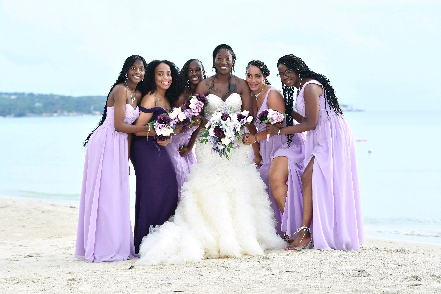 Bride with bridesmaids on a beach; bride in white, bridesmaids in purple, smiling, holding bouquets.