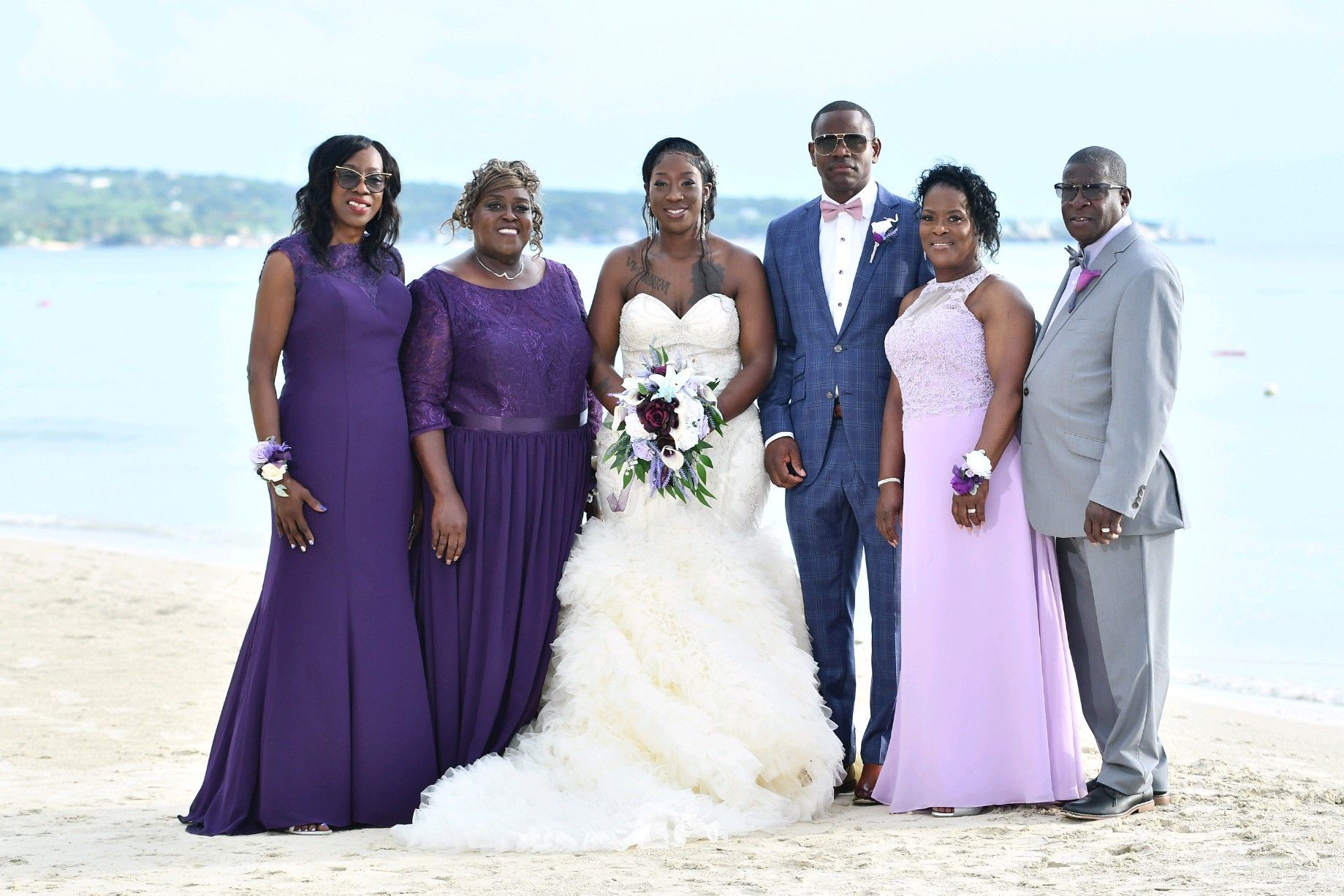 Wedding party on beach: Bride in white dress, groom in blue suit, family in purple.