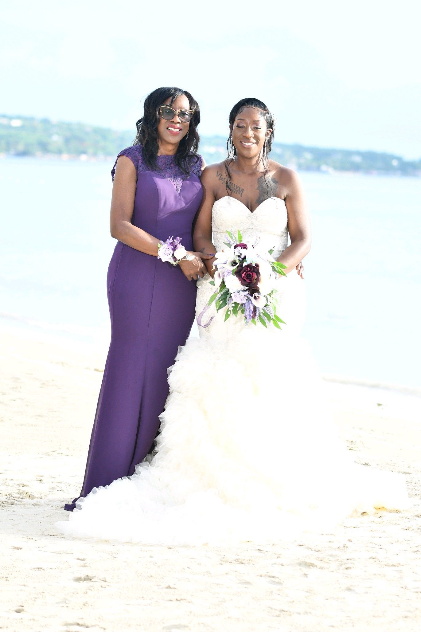 Bride in white dress and bridesmaid in purple dress standing on a beach.