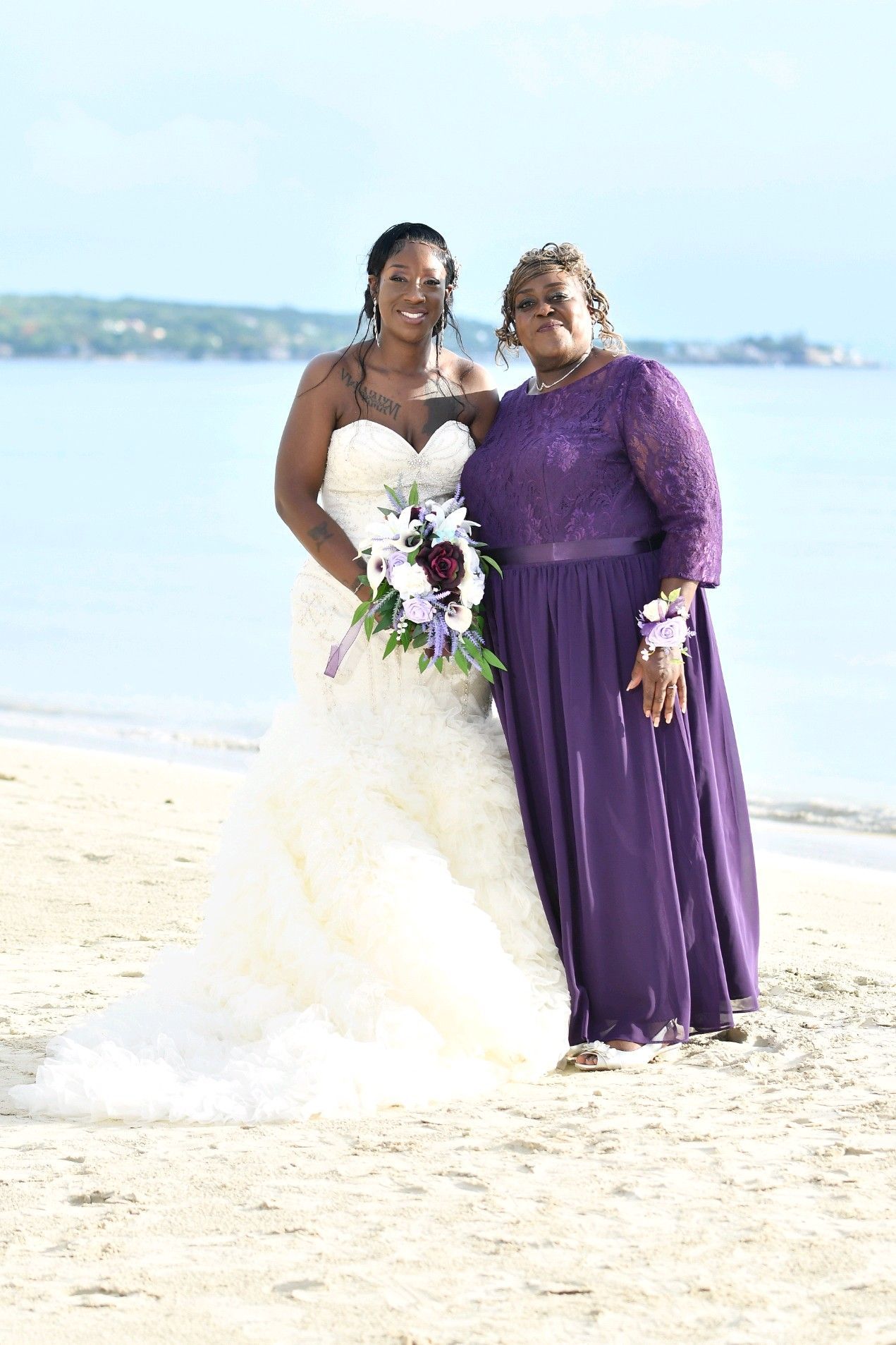 Bride in white gown with mother in purple dress, standing on a beach.