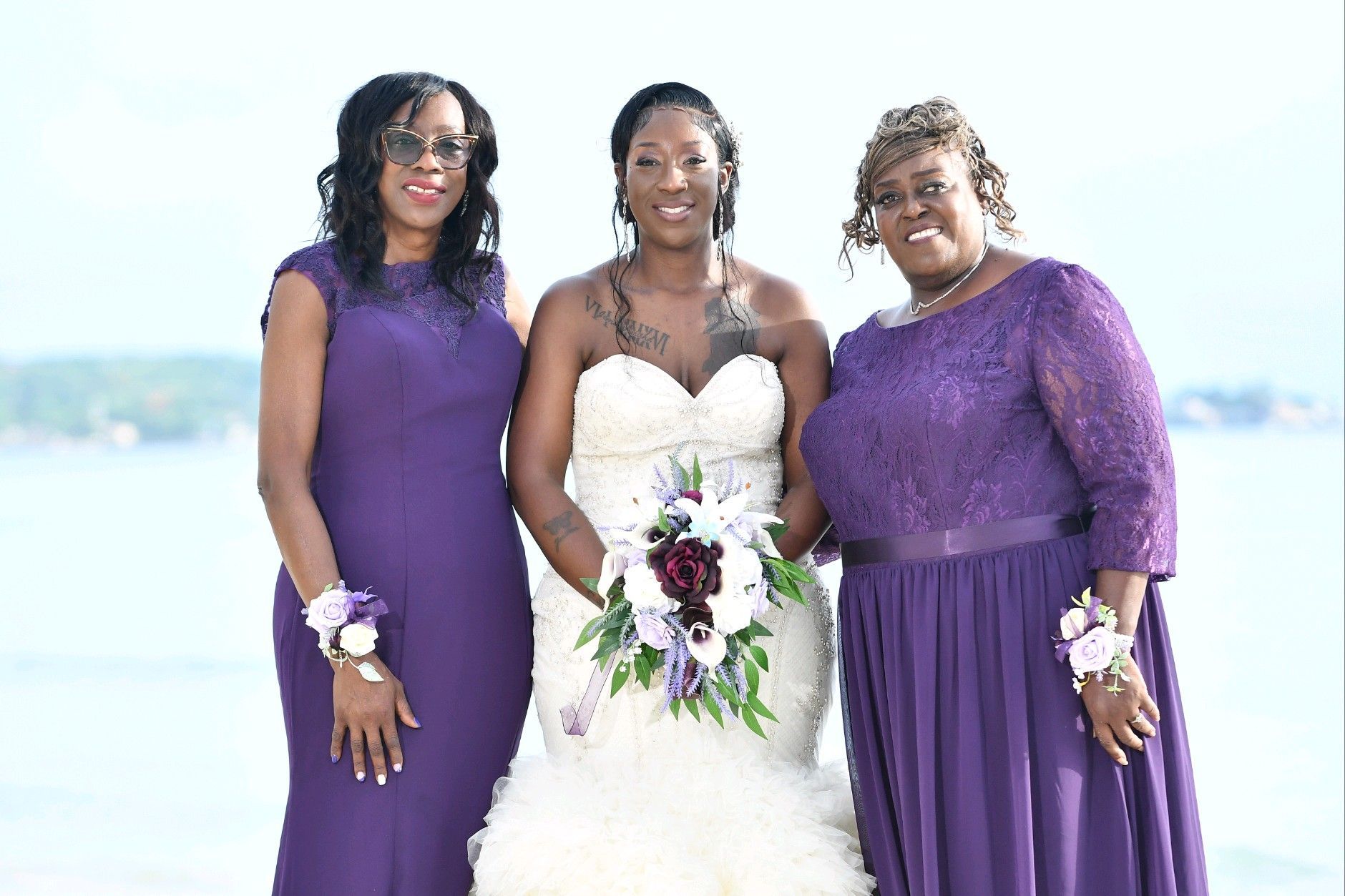 Bride with two women, all smiling, wearing purple dresses, posing by water.
