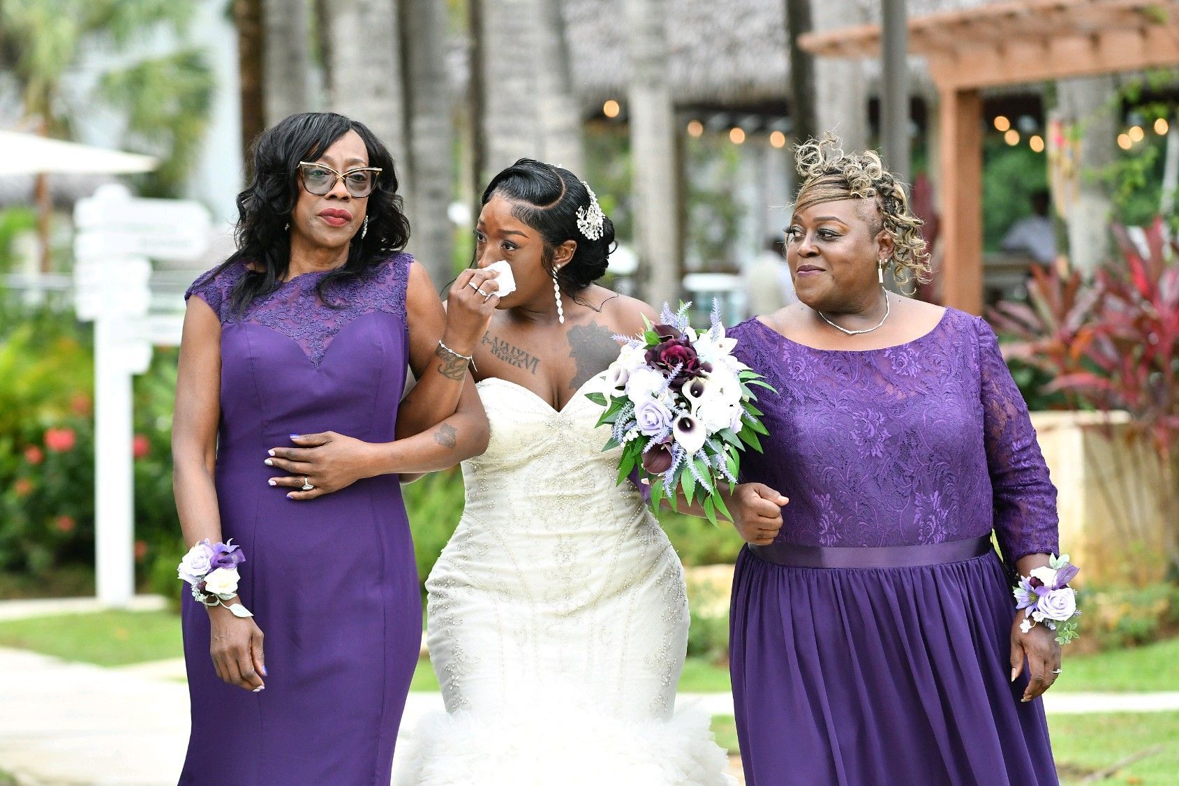 Bride crying, escorted by two women in purple dresses, walking outdoors at a wedding.
