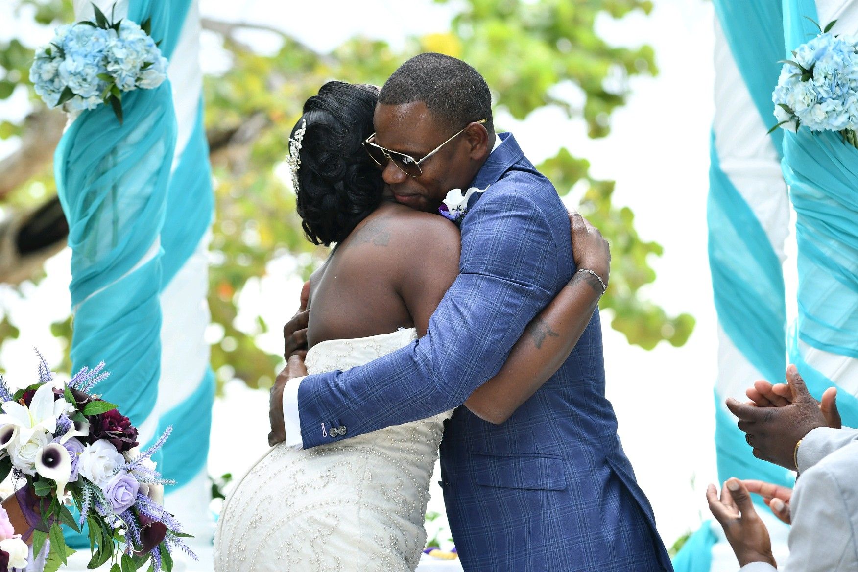 Newlyweds embrace during outdoor ceremony, teal and white decorations.